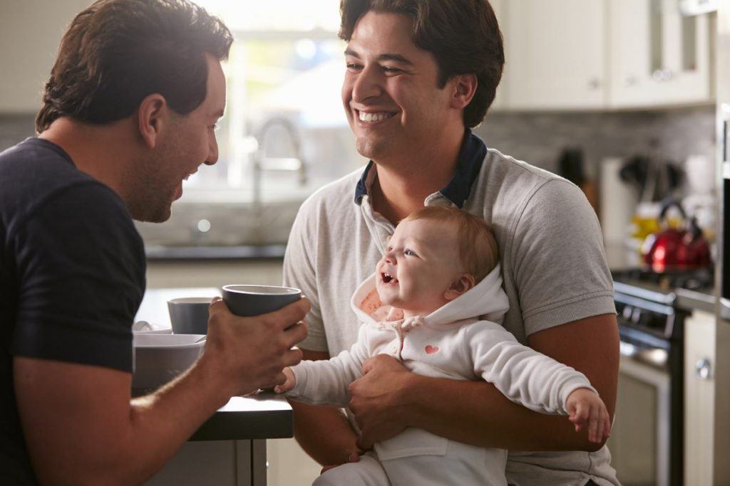 Male gay couple holding baby girl in their kitchen.